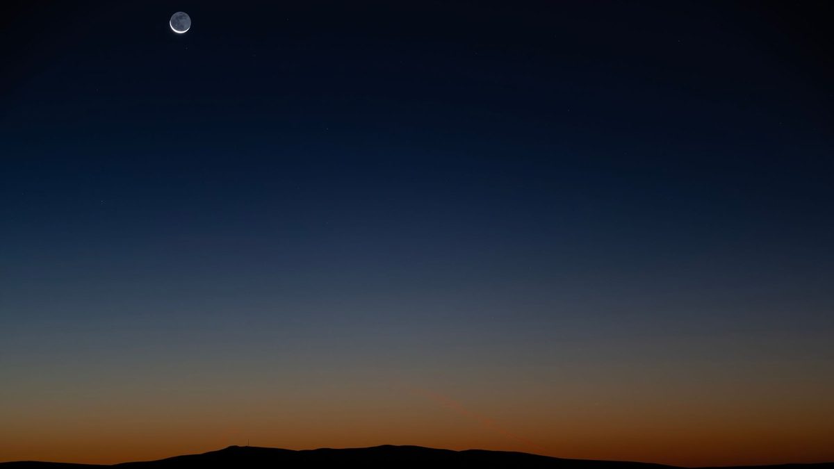 A thin crescent moon in a dark blue sky above a low hill line at dusk